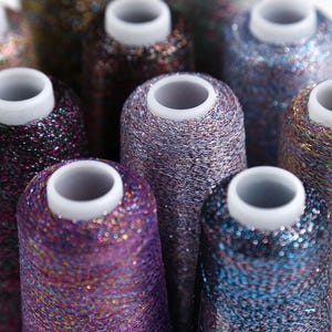 May include: Close-up of several spools of glittery thread in various colors, including purple, blue, and bronze. Each spool has a white core and is tightly wound with shimmering thread, creating a textured surface. The threads catch the light, giving them a sparkling appearance.