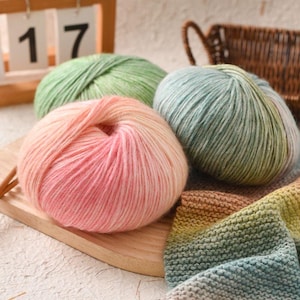 Puede incluir: Primer plano de cuatro ovillos de lana en tonos rosa, verde y azul. La lana es suave y esponjosa, con un efecto degradado en el ovillo rosa. Un calendario de madera con el número 17 está en el fondo. Ideal para proyectos de punto y ganchillo.
