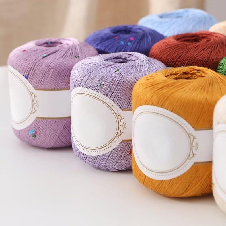 Puede incluir: Una colecci&oacute;n de madejas de hilo coloridas en tonos morado, naranja, rojo y azul. Cada madeja tiene una etiqueta blanca en blanco con un borde adornado. El hilo est&aacute; bien enrollado, lo que sugiere que est&aacute; listo para proyectos de tejido o ganchillo.