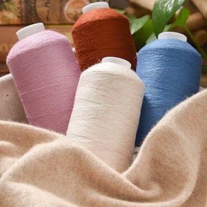 Könnte beinhalten: Nahaufnahme von Garnrollen in Rosa, Orange, Weiß und Blau. Die Spulen haben weiße Kappen und liegen auf einem beigen Stoff, passend für Bastel- oder Textilprojekte.