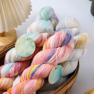 May include: A collection of colourful yarn skeins in a basket. The yarn features shades of pink, blue, yellow, and white, giving a pastel appearance. Each skein has a tag attached. The yarn is suitable for knitting or crochet projects.