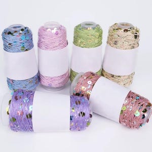 Peut inclure: Plusieurs bobines de fil coloré avec des paillettes, dans des tons de bleu, rose, vert et or. Chaque bobine est enveloppée d'une étiquette blanche. Idéal pour les projets de couture et de loisirs créatifs.