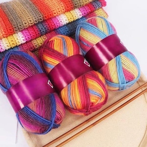 Op de afbeelding: Verschillende bollen veelkleurig garen in levendige tinten roze, blauw, geel en oranje, omwikkeld met een dieppaarse band. Breinaalden en een opgevouwen gebreide deken zijn ook zichtbaar, wat wijst op een knutselthema.