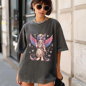 May include: A charcoal grey short-sleeve t-shirt featuring a graphic print of a small figure with colourful wings, wearing sunglasses and a fringed outfit, surrounded by feathers, small geometric patterns, and two bull skull icons.