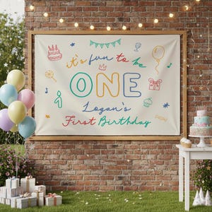 May include: A personalized birthday banner with the text "it's fun to be ONE" and "Logan's First Birthday" in colorful lettering. The banner is decorated with festive illustrations and is displayed on a brick wall, with balloons and gifts nearby.