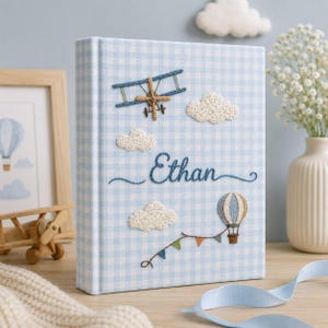 May include: A light blue and white gingham photo album with embroidered details. The cover features a biplane, clouds, a hot air balloon, and the name "Ethan" in cursive. A decorative item for baby photos.