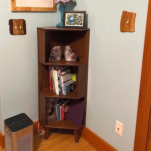 May include: A dark brown corner bookshelf with three shelves, holding books, amethyst stones, and a purple lamp. A framed picture of a bird and a small heater are also visible. Wooden light switch covers are mounted on the wall.
