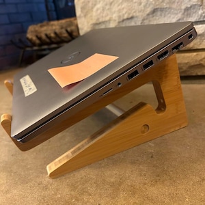 May include: A silver laptop computer rests on a natural bamboo laptop stand. The stand is angled for ergonomic use. A peach-colored sticky note is affixed to the laptop's surface. The background features a stone fireplace.