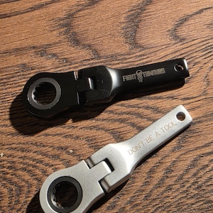 May include: Two ratcheting spanners, one black and one silver, are on a wooden surface. The black spanner has the text "FIGHT TUMBLERS" and a skull logo. The silver spanner has the text "DON'T BE A TOOL".