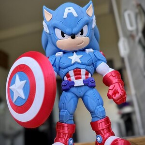 Op de afbeelding: Een blauw en rood actiefiguur van een Sonic the Hedgehog-Captain America hybride. Het figuur houdt een rood, wit en blauw schild vast. Het figuur draagt een blauw pak met een witte ster en rode laarzen.