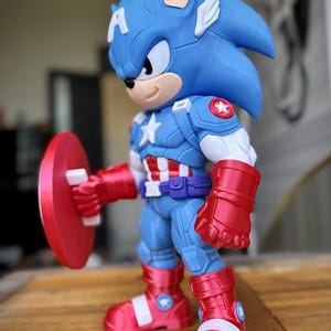 Op de afbeelding: Een blauw en rood beeldje van een personage dat een hybride is van Captain America en Sonic the Hedgehog. Het beeldje draagt een Captain America-pak en houdt een rood schild vast. Het beeldje heeft een blauwe kop met witte oren en een witte ster op de borst.