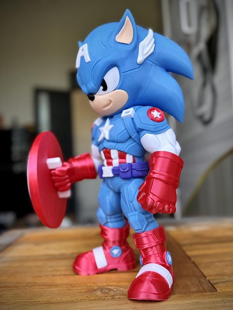Op de afbeelding: Een blauw en rood beeldje van een personage dat een hybride is van Captain America en Sonic the Hedgehog. Het beeldje draagt een Captain America-pak en houdt een rood schild vast. Het beeldje heeft een blauwe kop met witte oren en een witte ster op de borst.