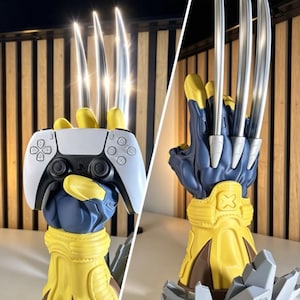 May include: A blue and yellow gauntlet stand featuring three long, metallic silver claws extending from the knuckles. The gauntlet is designed to hold a white and black game controller, with a base shaped like jagged gray rock fragments.