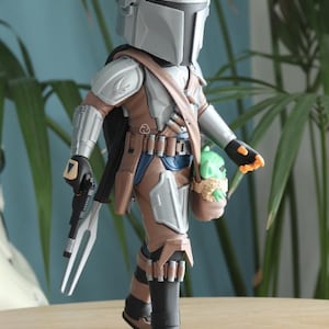 May include: A detailed action figure of The Mandalorian, featuring a silver helmet and armor, brown and blue clothing, and a black cape. The figure carries a blaster and a small green Grogu figure in a satchel. The figure is approximately 6 inches tall.
