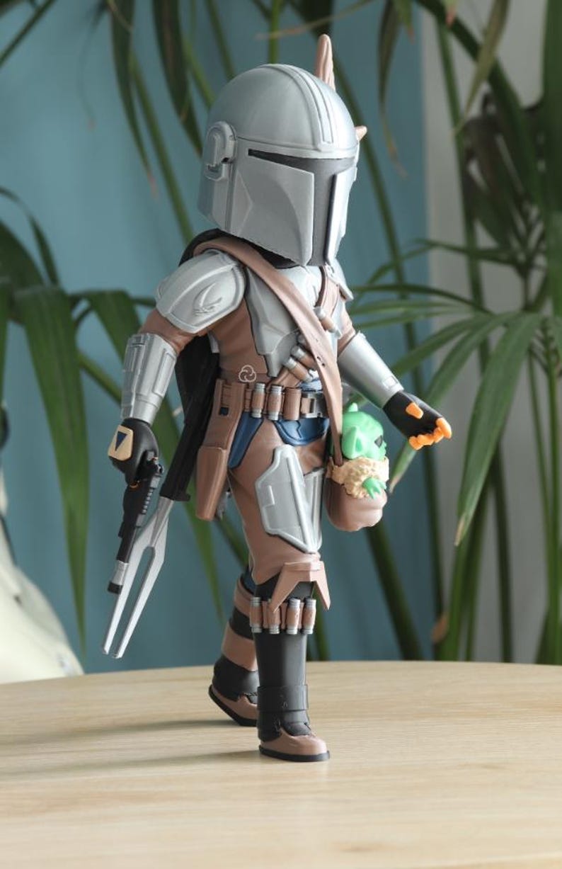 May include: A detailed action figure of The Mandalorian, featuring a silver helmet and armor, brown and blue clothing, and a black cape. The figure carries a blaster and a small green Grogu figure in a satchel. The figure is approximately 6 inches tall.