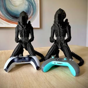 May include: Two black 3D-printed Xenomorph figures in a kneeling position, each designed to hold a gaming controller. One figure holds a white and black controller, while the other holds a gray and light blue controller, both positioned upright.