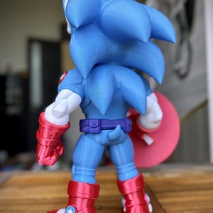 Op de afbeelding: Een blauw en rood actiefiguur van Sonic the Hedgehog in een op Captain America ge&iuml;nspireerde outfit. Het figuur heeft een blauw lichaam, rode laarzen en een rood schild. Het figuur staat op een houten oppervlak.