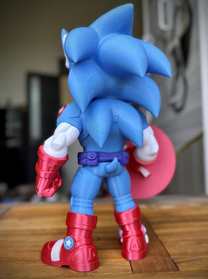 Op de afbeelding: Een blauw en rood actiefiguur van Sonic the Hedgehog in een op Captain America ge&iuml;nspireerde outfit. Het figuur heeft een blauw lichaam, rode laarzen en een rood schild. Het figuur staat op een houten oppervlak.