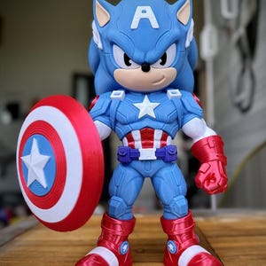Op de afbeelding: Een blauw en rood beeldje van Sonic the Hedgehog verkleed als Captain America, met een schild. Het beeldje heeft een blauw lichaam, rode laarzen en handschoenen en een witte ster op de borst. Het schild is rood, wit en blauw.