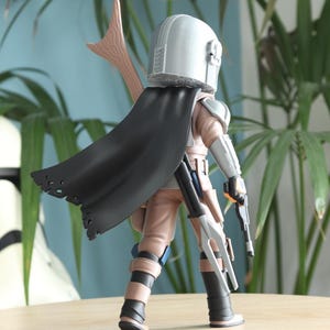 May include: A detailed action figure of a character from a science fiction franchise. The figure is wearing a silver helmet, a black cape, and tan and gray armor. It is holding two blasters and a spear-like weapon.