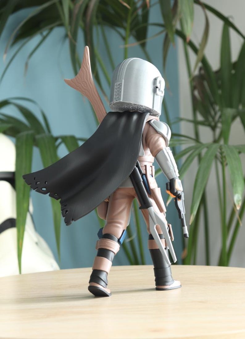 May include: A detailed action figure of a character from a science fiction franchise. The figure is wearing a silver helmet, a black cape, and tan and gray armor. It is holding two blasters and a spear-like weapon.