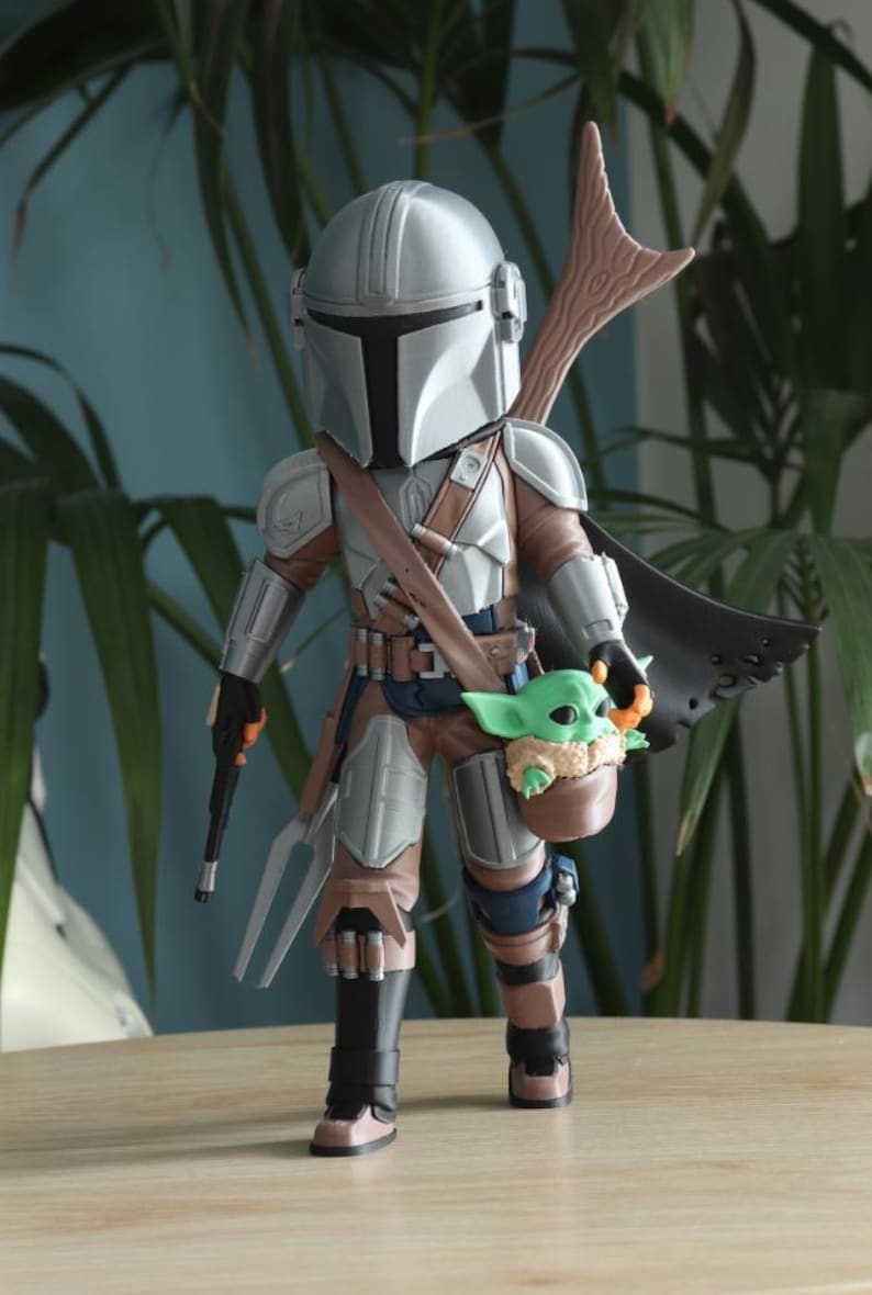 May include: A detailed figurine of The Mandalorian, clad in silver and brown armor, holding a weapon and a small green creature. The figure has a black cape and a wooden spear-like attachment. The figurine is standing on a wooden surface.