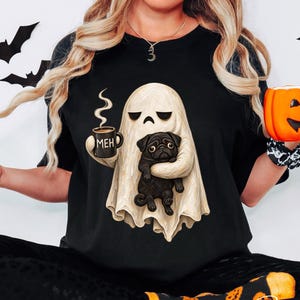 May include: A black t-shirt featuring a graphic of a white ghost holding a black pug and a black coffee mug with the text MEH printed on it. The ghost has a neutral expression. The shirt is worn with black patterned leggings and orange socks.