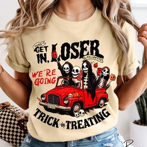 May include: A light yellow t-shirt featuring a graphic of four skeletons in a red vintage car. The text reads: Happy Get In, Loser We're Going Trick Or Treating. The skeletons hold red cups and pumpkins, with a small skull on the car hood.