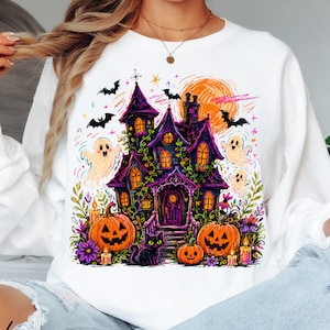 May include: A white long-sleeve sweatshirt featuring a colourful graphic print of a haunted house, jack-o'-lanterns, a black cat, ghosts, and bats. The design includes vibrant purple, orange, and yellow tones with stars and floral accents.