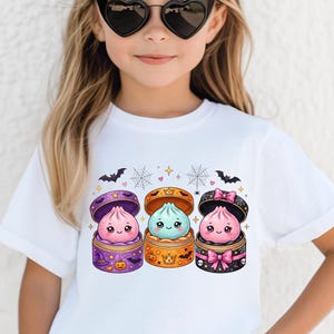 Könnte beinhalten: Ein weißes T-Shirt mit einer Grafik von drei niedlichen, lächelnden Teigtaschen in offenen Bambusdämpfern. Die Dämpfer sind mit Halloween-Motiven wie Fledermäusen, Spinnennetzen, Kürbissen und Kronen in Lila, Orange und Schwarz verziert.