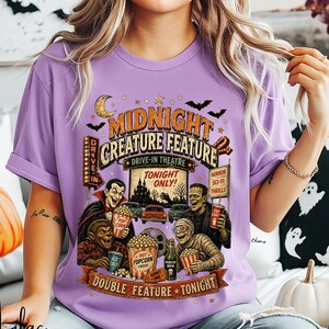 Pode incluir: Uma t-shirt roxo-claro com uma estampa gráfica de uma cena de cinema drive-in com personagens clássicos de terror, pipocas e o texto "MIDNIGHT CREATURE FEATURE", "TONIGHT ONLY", "DRIVE-IN", "HORROR SCI-FI THRILLS" e "DOUBLE FEATURE TONIGHT".