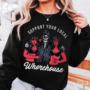 May include: A black long-sleeve sweatshirt featuring a graphic print of a central skeleton figure in a dark hooded robe flanked by two red-skinned figures with horns. The text "SUPPORT YOUR LOCAL" appears above and "WHOREHOUSE" below.