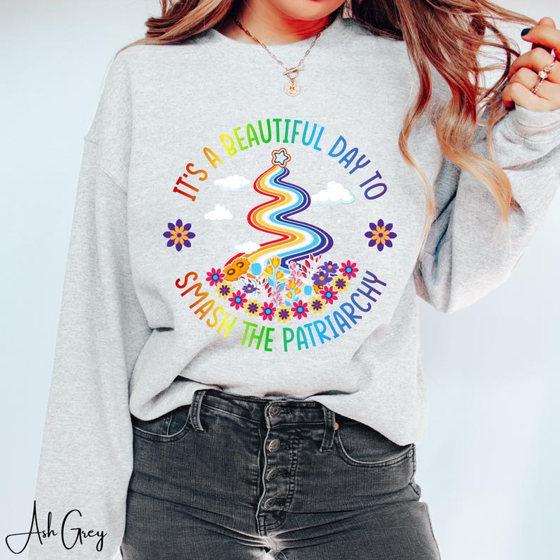 Op de afbeelding: Lichtgrijze sweatshirt met een kleurrijke afbeelding met de tekst "It's a beautiful day to smash the patriarchy." Het ontwerp bevat een regenboog, bloemen en tekst in een cirkelvormige opstelling. De sweatshirt is een casual stijl.