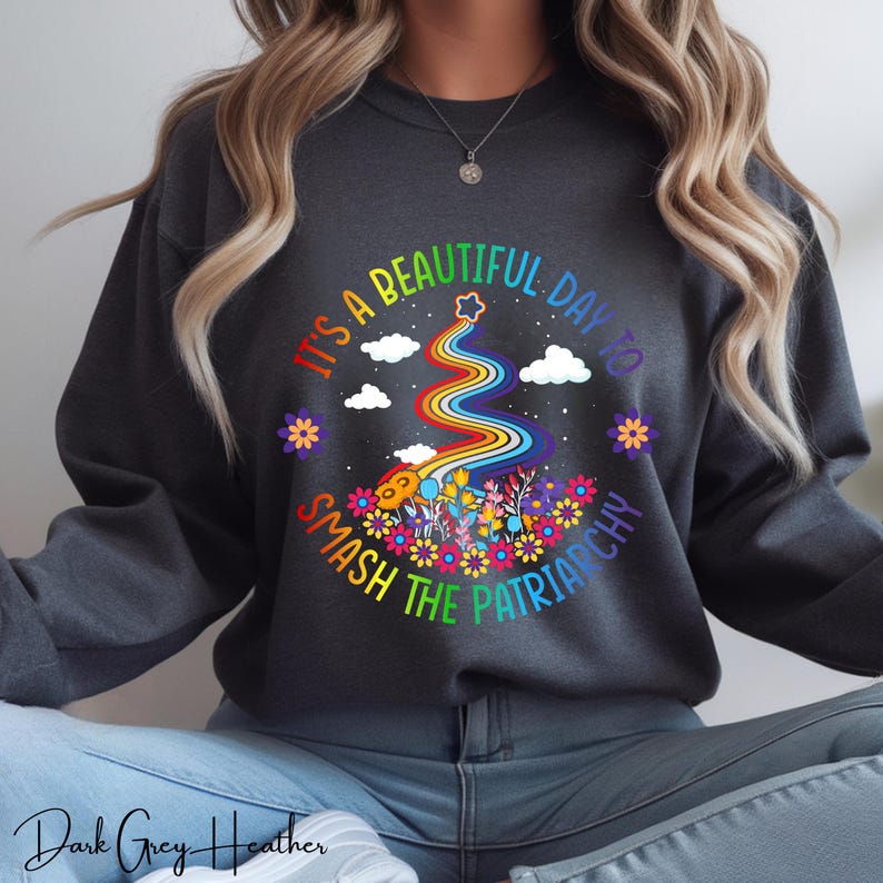 Op de afbeelding: Donkergrijze heather sweatshirt met de tekst "IT'S A BEAUTIFUL DAY TO SMASH THE PATRIARCHY". Het ontwerp toont een regenboog, bloemen, wolken en een ster. De sweatshirt heeft een ronde hals.