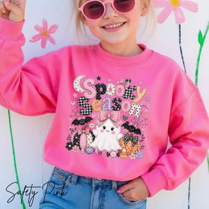 May include: A vibrant pink sweatshirt featuring a Halloween design. The graphic reads "Spooky Season" in a patchwork style, with a friendly ghost, pumpkins, bats, and spiderwebs. The sweatshirt is worn by a person wearing pink sunglasses.