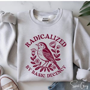 以下が含まれることがあります： バーガンディ色の「RADICALIZED BY BASIC DECENCY」の文字が入ったライトグレーのスウェットシャツ。バーガンディ色の鳥のイラストが花のリースの中に描かれています。スウェットシャツはスポーツグレーの色です。