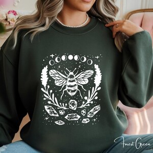 May include: Forest green sweatshirt featuring a white graphic of a bee, crystals, moon phases, and botanical elements. The design is centered on the front of the sweatshirt. The words "Forest Green" are written in cursive on the bottom right.