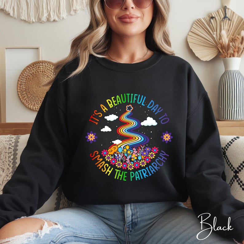 Op de afbeelding: Zwart sweatshirt met een kleurrijke afbeelding met de tekst "It's a beautiful day to smash the patriarchy". Het ontwerp bevat een regenboog, bloemen, wolken en sterren. Het woord "Black" staat cursief rechtsonder.