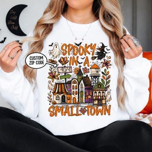 May include: A white long-sleeve sweatshirt featuring a colorful graphic print of a Halloween-themed town with ghosts, a witch, and a sign reading 4074. The text SPOOKY IN A SMALL TOWN is printed in orange above and below the illustrated buildings.