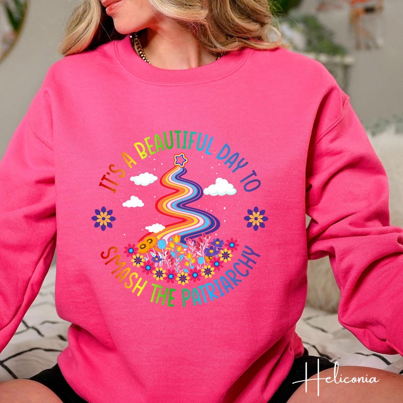 Op de afbeelding: Een felroze sweatshirt met een kleurrijke afbeelding met de tekst "It's a beautiful day to smash the patriarchy." De afbeelding bevat een regenboog, wolken, bloemen en een ster. De sweatshirt heeft een ronde hals.