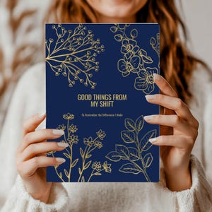 May include: A navy blue notebook with gold floral illustrations and the text "GOOD THINGS FROM MY SHIFT" and "To Remember the Difference I Made". The notebook is held by a person wearing a white sweater.