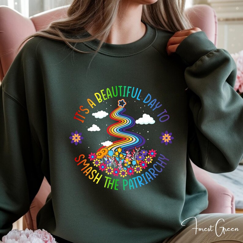 Op de afbeelding: Bost groene sweatshirt met een kleurrijke afbeelding. De afbeelding toont een regenboog, bloemen, wolken en de tekst "It's a beautiful day to smash the patriarchy."
