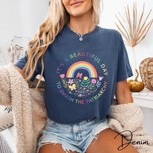 Op de afbeelding: Marineblauwe t-shirt met een regenboog, bloemen en vlinders. De tekst luidt: "It's a beautiful day to smash the patriarchy." Een casual, comfortabele stijl.
