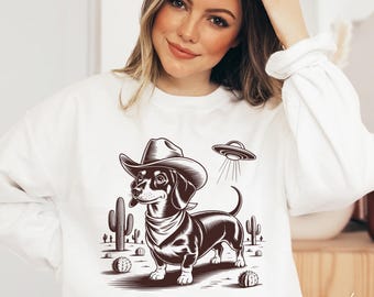 May include: White sweatshirt featuring a cartoon dachshund wearing a cowboy hat and bandana, with a UFO above. The design includes cacti and desert elements. The word "White" is printed in the bottom right corner.