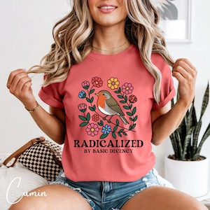 Puede incluir: Camiseta de color coral con una ilustración de un pájaro rodeado de flores. El pájaro tiene el pecho rojo, alas grises y vientre blanco. El texto "RADICALIZED BY BASIC DECENCY" está impreso debajo del diseño del pájaro.