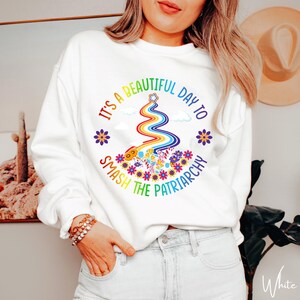 Op de afbeelding: Witte sweatshirt met een regenboogontwerp en de tekst "It's a beautiful day to smash the patriarchy." Het ontwerp bevat een regenboog, bloemen en een ster. De sweatshirt heeft een ronde hals.