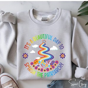 Op de afbeelding: Een lichtgrijs sweatshirt met de tekst "IT'S A BEAUTIFUL DAY TO SMASH THE PATRIARCHY" in regenboogkleuren. Het ontwerp bevat een regenboog, bloemen en wolken. Het sweatshirt is gelabeld "Sport Grey".