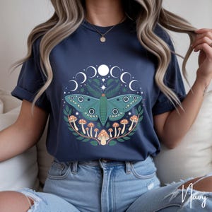 May include: Navy blue t-shirt featuring a teal moth design surrounded by a crescent moon and mushroom graphic. The moth has detailed wings and is centered on the shirt. The design is encircled by a wreath of leaves and mushrooms.