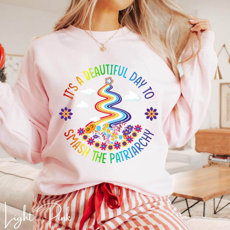 Op de afbeelding: Lichtroze sweatshirt met de tekst "It's a beautiful day to smash the patriarchy" in een cirkelvormig ontwerp. Het ontwerp bevat een regenboog, bloemen en een ster. De woorden "Light Pink" staan onderaan.