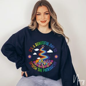 Op de afbeelding: Marineblauwe sweatshirt met een regenboogontwerp en de tekst "It's a beautiful day to smash the patriarchy." De afbeelding bevat een regenboog, bloemen, wolken en een ster.